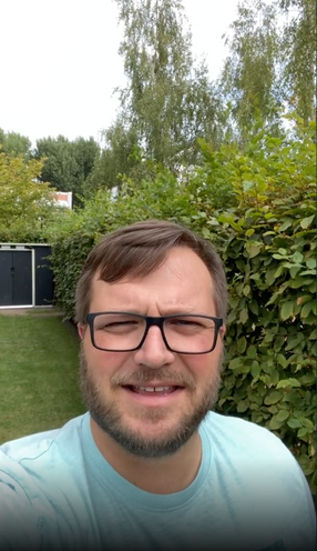 Mann im Garten vor Hecke und Rasen spricht in die Kamera