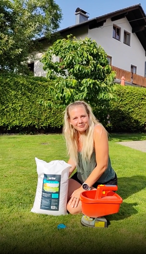 Frau kniet im Garten neben Sack turbogrün Sommerrasendünger und orangem Streuer
